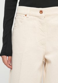 Άτομο που φοράει ψηλόμεσο εκρού τζην παντελόνι με χάλκινους κουμπιά και μαύρη ριμπ μακρυμάνικη μπλούζα, με το χέρι να ακουμπάει στο πλάι.