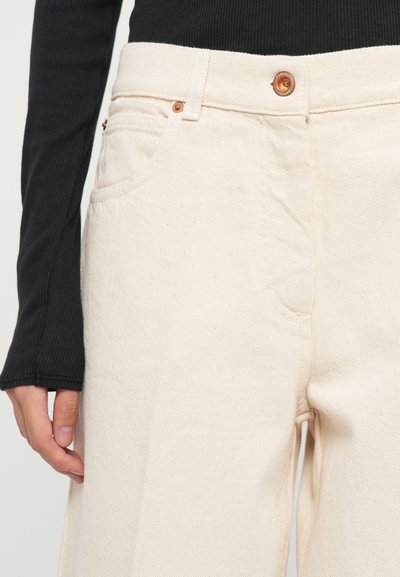 Persoană îmbrăcată cu pantaloni din denim albaștri cu talie înaltă, prevăzuți cu nasturi din cupru, și un top lung din material ribbed de culoare neagră, cu mâna odihnindu-se pe lateral.