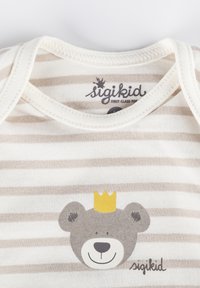 Gestreifte beige und weiße Stoffbluse mit einer grauen Bärengrafik und einer gelben Krone. Das Etikett trägt die Aufschrift "sigikid" in einer verspielten Schriftart.