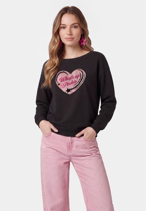Ung kvinde med langt hår iført sort sweatshirt med lyserødt hjerte og tekst "What's up Pookie" og lyserøde bukser, poserende med hænderne i lommerne.