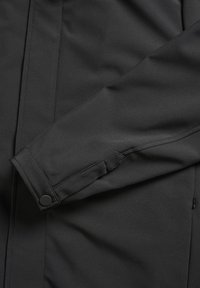 Manche de veste noire avec poignet boutonné et lignes de couture visibles sur une surface de tissu lisse.