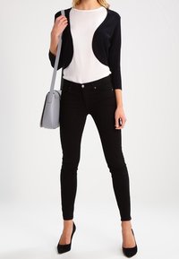 Zwarte slim fit jeans gecombineerd met een witte top en een zwart cropped cardigan. Accessoires bestaan uit een lichtgrijze handtas en zwarte hakken.
