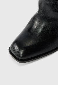 Close-up of glossy black square-toe leather boot with smooth textured surface against white background.