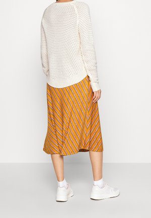 Personne portant un pull en maille blanc, une jupe midi rayée orange, des chaussettes blanches et des baskets blanches, debout de dos sur un fond uni.