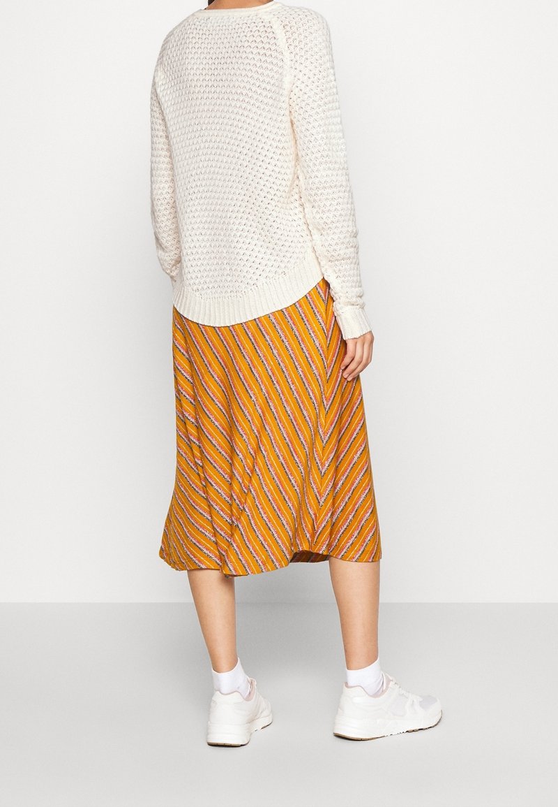 Personne portant un pull en maille blanc, une jupe midi rayée orange, des chaussettes blanches et des baskets blanches, debout de dos sur un fond uni.