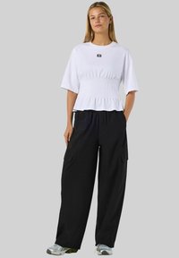 Bijela majica kratkih rukava s detaljem skupljenog struka, uparena s labavim crnim cargo hlačama. Model nosi sive tenisice. Jednostavni logo na majici.