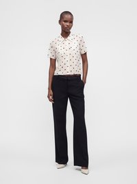 Femme portant un chemisier blanc à manches courtes avec des pois marron, un pantalon noir à jambes larges et des chaussures blanches à bouts pointus, debout devant un fond uni.