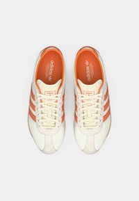 Λευκά και πορτοκαλί αθλητικά παπούτσια με τρεις πορτοκαλί λωρίδες. Υφή στη γλώσσα, ελαφριές κορδόνια, ορατό λογότυπο στο εσωτερικό. Επίπεδη ελαστική σόλα.