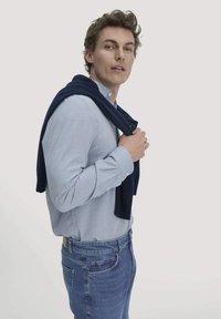 Jonge man in een lichtblauw shirt en blauwe spijkerbroek, met een marineblauwe trui over zijn schouders, tegen een effen lichte achtergrond.