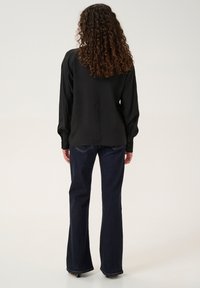 Blusa nera a maniche lunghe con tessuto strutturato; abbinata a jeans svasati blu scuro aderenti. La blusa presenta polsini con bottone e una linea dritta.