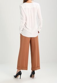 Chemise blanche à manches longues avec un col rond et une couture dorsale subtile, associée à un pantalon taille haute, jambes larges, de couleur marron, et des talons noirs.