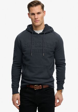Mand iført en mørkeblå hættetrøje med en frontlomme, brun bælte, sorte bukser og et armbåndsur, stående mod en ensartet hvid baggrund.