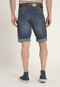 Shorts di jeans blu scuro, risvolti, design a cinque tasche, cintura in pelle marrone, abbinati a sneakers alte navy. Vista da dietro.