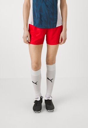 Rode sportshorts met een gestroomlijnd ontwerp, gecombineerd met grijze kniekousen met een zwart logo en zwarte voetbalschoenen.
