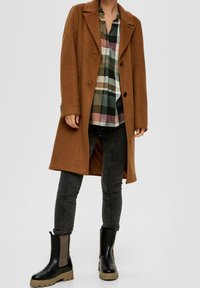 Cappotto di lana marrone con vestibilità sartoriale, ampi revers e due bottoni, indossato sopra una camicia a scacchi. Stivaletti neri alla caviglia con dettagli testurizzati.