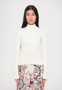 Krem pulover s turtleneck iz teksturirane tkanine in dvignjenim logotipom, v kombinaciji s hlačami s cvetličnim vzorcem v živahnih barvah.