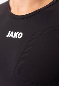Schwarzes Sportshirt mit kurzen Ärmeln, das mit strukturierten Akzenten und einem weißen Logo "JAKO" auf der Vorderseite auffällig versehen ist.