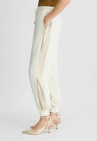Pantalon de jogging blanc avec une bande latérale en sequins dorés brillants, une taille élastique et des poignets. Assorti avec des escarpins à bride arrière dorés.