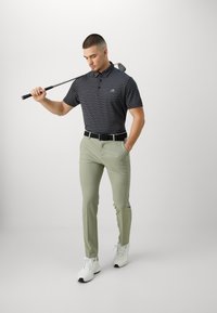 Πουκάμισο πόλο με ασπρόμαυρες ρίγες, ανοιχτοπράσινο παντελόνι, μαύρη ζώνη. Το μοντέλο κρατ�άει ένα γκολφ ρόπαλο, φοράει λευκά αθλητικά παπούτσια.