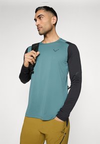 Türkisfarbenes Langarm-Athletikshirt mit schwarzen Akzenten. Verfügt über einen Rundhalsausschnitt und ein Logo auf der Brust. Leichtes, strukturiertes Gewebe, geeignet für sportliche Aktivitäten.