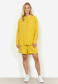 Gelbes Hemd mit Knopfleiste und Brusttasche, kombiniert mit gelben Shorts. Getragen mit weißen Sneakers. Glatter Stoff und entspannte Passform.