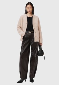 Kobieta w beżowej, puszystej kardiganie noszącym się na czarną bluzkę, czarnych szerokich skórzanych spodniach, czarnych botkach na obcasie, trzymająca małą czarną torebkę.