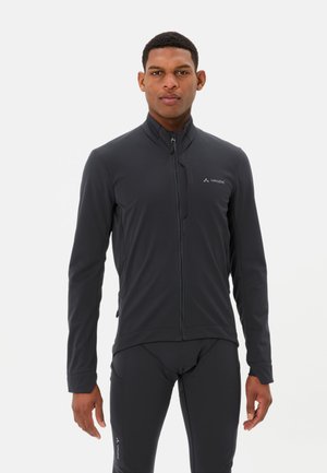 Mann trägt eine schwarze Vaude Zip-Up-Jacke und passende schwarze Hose, steht vor einem schlichten hellen Hintergrund.