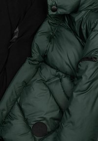 Giacca trapuntata verde scuro con tasca, zip e una toppa circolare nera accanto a un materiale tessile nero.