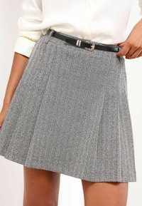 Harmaa houndstooth-kuvioinen hame, jossa on laskoksia. Varustettu mustalla vyöllä, jossa on hopeinen solki. Valmistettu teksturoidusta kankaasta.