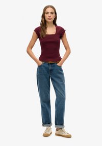 Burgunderfarbenes, figurbetontes Oberteil mit quadratischem Ausschnitt und fransigem Saum, kombiniert mit hochgeschnittenen blauen Jeans und beigen Sneakern.