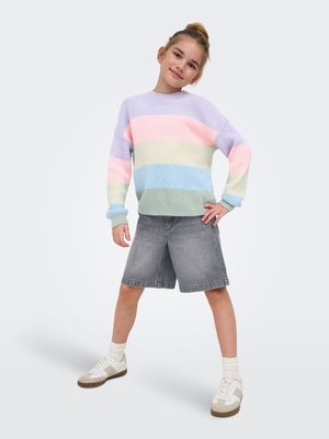 Ung pige med håret i knold iført pastelstribet sweater, grå knælange shorts, hvide sokker og sneakers, stående med hånden på hoften.