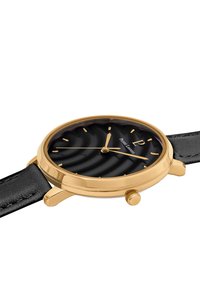 Guldfärgad armbandsklocka med en texturerad svart urtavla, guldfärgade timmarkörer och smal svart läderrem. Har en rund klocka och krona.