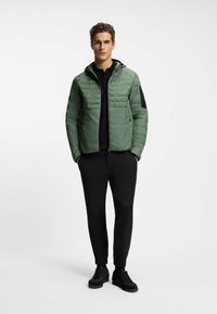Giovane uomo con capelli ricci che indossa una giacca verde imbottita, una camicia nera, pantaloni neri e sneakers nere, in piedi con le mani in tasca.