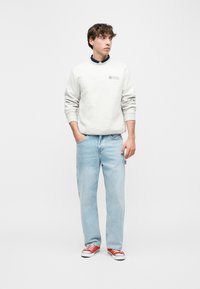 Νεαρός άνδρας στέκεται με τα χέρια στις τσέπες, φοράει λευκό φούτερ, ανοιχτόχρωμο τζιν και κόκκινα αθλητικά παπούτσια, κοιτάει προς τα δεξιά σε απλό φόντο.