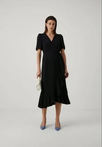 Vestido preto envelope com mangas curtas bufantes, decote em V, bainha rodada e detalhe de laço lateral. Usado com sapatos de salto azul claro e uma pequena bolsa branca.
