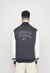 Musta-valkoinen Nike yliopistojakki, jossa on brodeerattu logo, neuloskaulus ja hihansuut, kontrastiväriset hihat ja teksturoitu kangas.