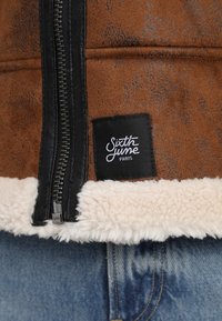 Brun faux læderjakke med sort lynlås, cremefarvet sherpa-foring og mærkat der viser "Sixth June Paris." Klassisk design med tekstureret detaljering.