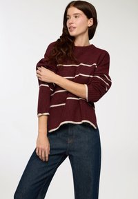 Kobieta w bordowym swetrze w paski i niebieskich dżinsach, z jednym ramieniem zgiętym w dół ciała, patrząca w bok na jednolitym tle.