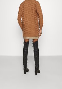 Persona vistiendo un suéter largo marrón texturizado, una falda mini beige y botas de tacón negras hasta la rodilla, de pie contra un fondo simple.