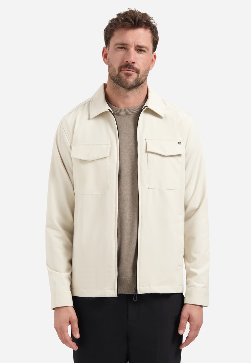 Man in een lichtbeige jasje over een taupe trui en zwarte broek, staand tegen een effen witte achtergrond.