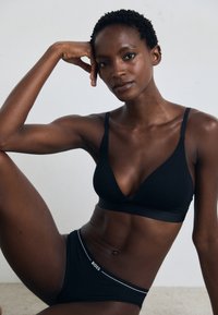 Soutien-gorge sans fil noir avec un design en V profond, associé à un slip noir assorti doté d'une ceinture avec logo. Tissu lisse et extensible.