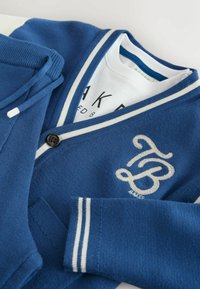 Modrá cardigan s bielymi pruhmi a gombíkovým zapínaním. Na prednej strane má grafické logo vyšívané striebornou niťou. Mäkká pletená textúra.