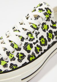 Białe trampki z czarno-neonowozielonym wzorem w panterkę, z białymi sznurowadłami, srebrnymi oczkami i czarną gumową podeszwą.