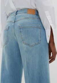 Jasnoniebieskie dżinsy z dwoma tylnymi kieszeniami i brązową skórzaną łatą, noszone z białą koszulą z guzikami, ręce luźno opadające po bokach.