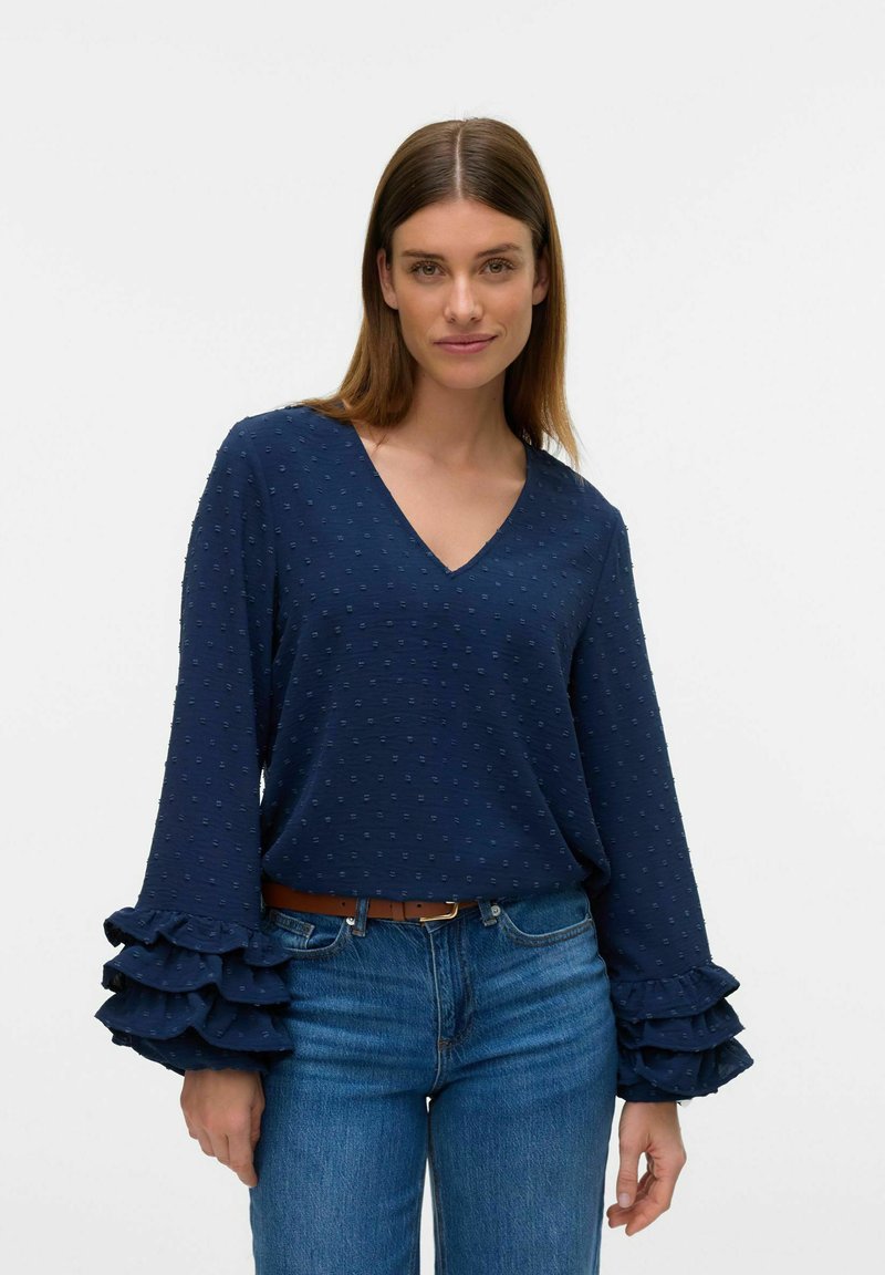 Blusa blu navy con scollo a V, decorata con pois testurizzati e maniche lunghe a volant, abbinata a jeans azzurri e una cintura marrone.