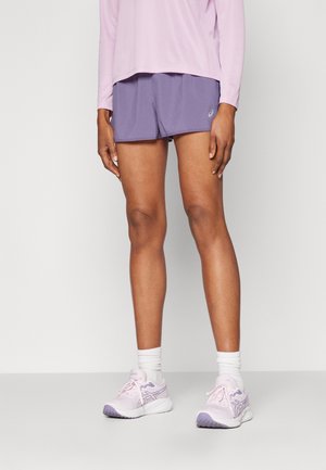 Person iført lyse lilla shorts, lyserød langærmet top, hvide sokker og lyserøde løbesko står på en almindelig gulv.