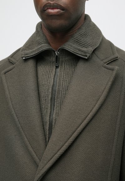 Olīvzaļš mētelis ar tekstūru apkakli un ar rāvējslēdzēju aizmugurē. Iezīmēts ar pielāgotām atloka malām un ribo detaļām iekšējā slānī.