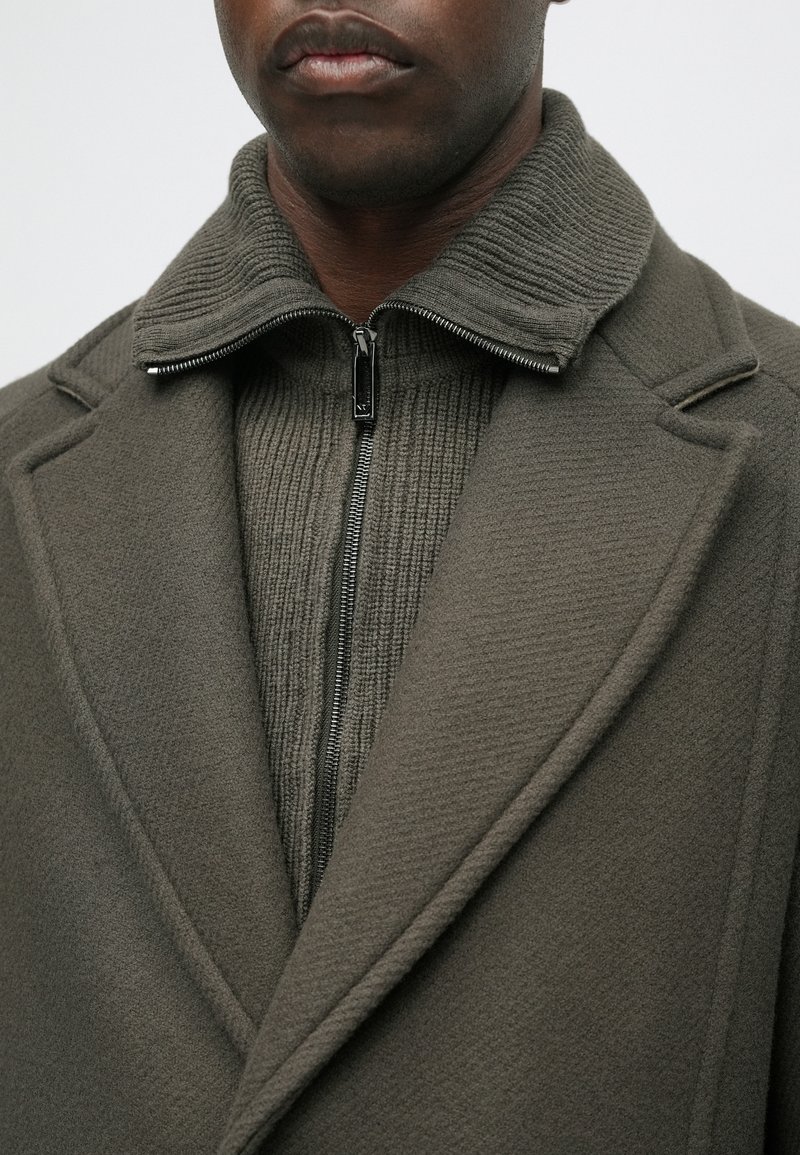 Olijfgroene jas met een getextureerde kraag en een rits aan de voorkant. Heeft op maat gemaakte revers en geribbelde details op de binnenlaag.