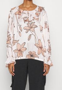 Blusa floreale di un rosa chiaro con motivi botanici in pesca e marrone, scollatura a volant e maniche elasticizzate, indossata con pantaloni neri.