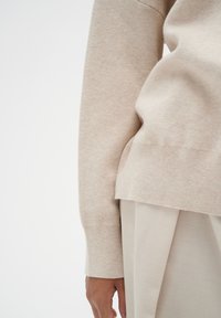 Beige knitted sweater featuring a crew neckline, ribbed cuffs, and a slightly loose fit; paired with light, pleated fabric trousers.